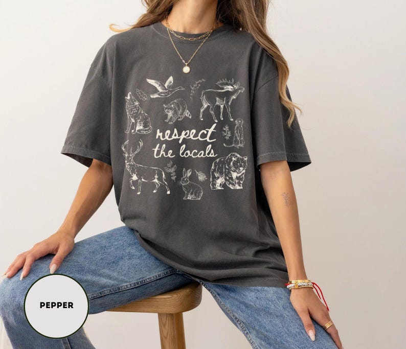 May include: Dark grey t-shirt with the text "respect the locals" and illustrations of various animals, including a wolf, deer, and bear. The shirt is worn by a person wearing blue jeans.