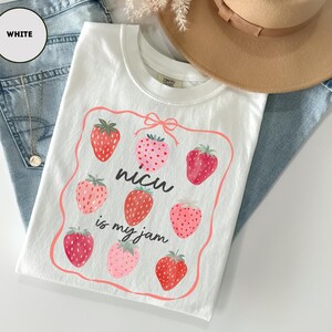 May include: White t-shirt with a strawberry design. The shirt features a rectangular frame with eight strawberries and the text "nicu is my jam". The shirt is laid on top of blue jeans and a tan hat.