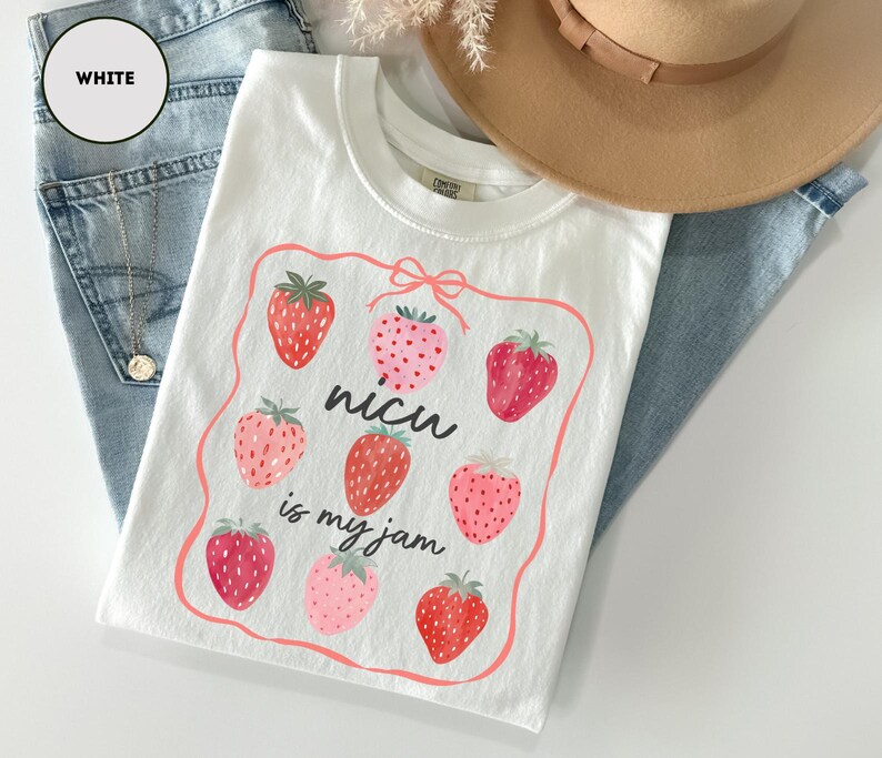 May include: White t-shirt with a strawberry design. The shirt features a rectangular frame with eight strawberries and the text "nicu is my jam". The shirt is laid on top of blue jeans and a tan hat.