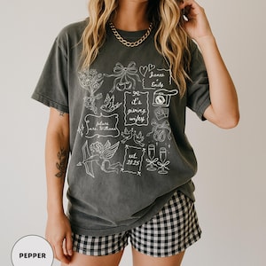 May include: A dark gray t-shirt with a white graphic design featuring floral elements, doves, and the text "it's giving wifey." The shirt also includes the names "James & Emily" and the year "2025."