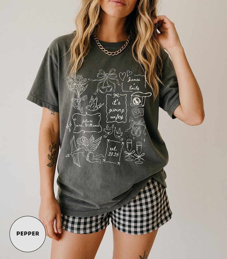 May include: A dark gray t-shirt with a white graphic design featuring floral elements, doves, and the text "it's giving wifey." The shirt also includes the names "James & Emily" and the year "2025."
