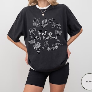 May include: Black t-shirt with white graphic illustrations of wedding-related items, including champagne glasses, shoes, and a ring. The text "Future Mrs. Williams" is centered on the front. The shirt is worn with black shorts.