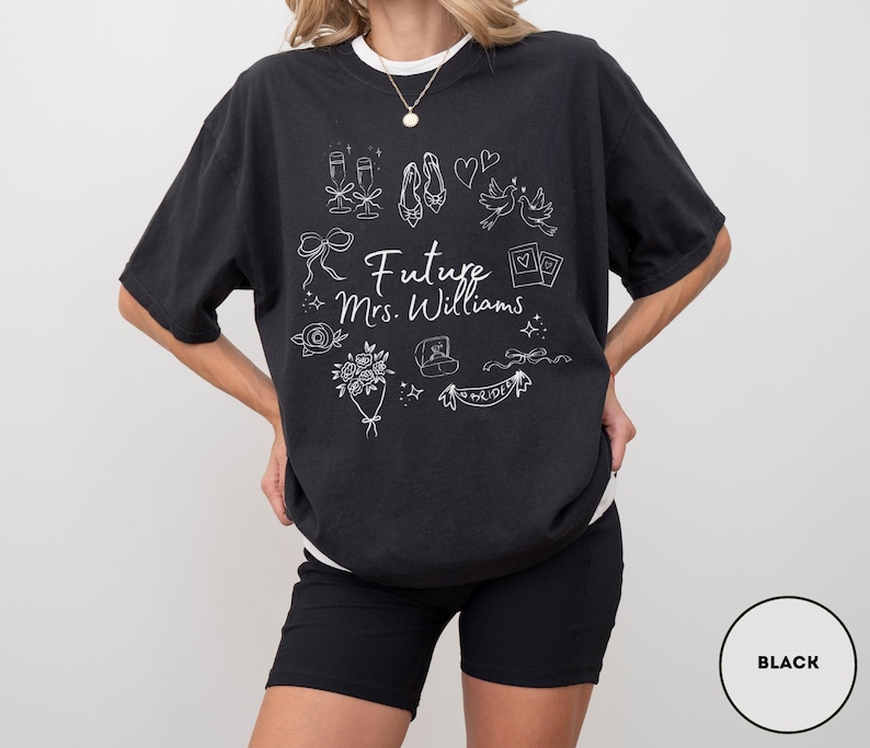 May include: Black t-shirt with white graphic illustrations of wedding-related items, including champagne glasses, shoes, and a ring. The text "Future Mrs. Williams" is centered on the front. The shirt is worn with black shorts.