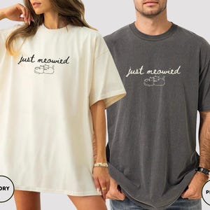 May include: Ivory and pepper-colored oversized t-shirts. Each shirt features the text "just meowied" in a script font, with a small illustration of two cats. The ivory shirt is worn by a person, and the pepper shirt is worn by another person.