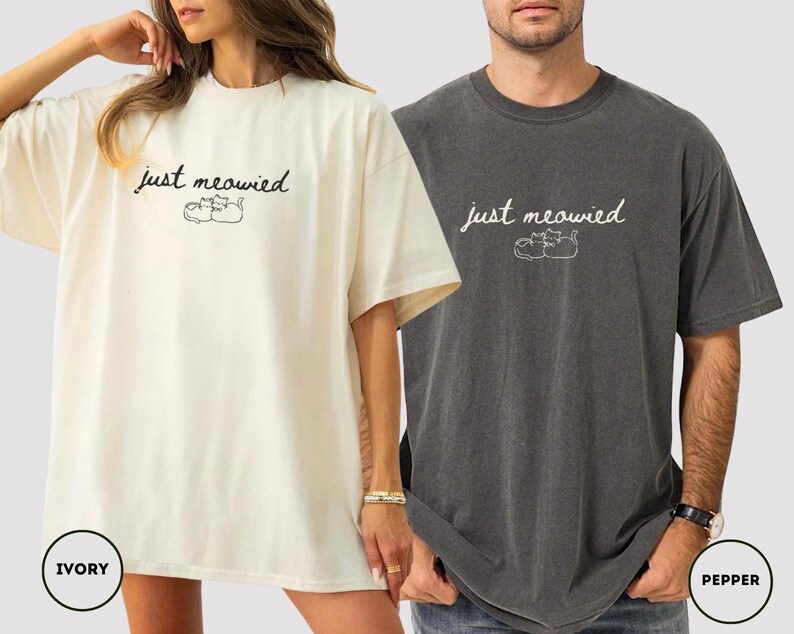 May include: Ivory and pepper-colored oversized t-shirts. Each shirt features the text "just meowied" in a script font, with a small illustration of two cats. The ivory shirt is worn by a person, and the pepper shirt is worn by another person.