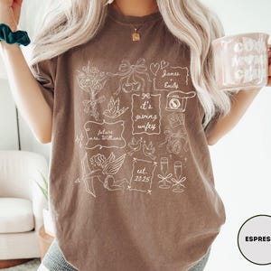 May include: Espresso-colored t-shirt with white line art illustrations, including flowers, doves, and the text "it's giving wifey." The shirt also features the text "future mrs. Williams" and "est. 2025."