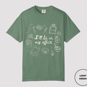 May include: Light green t-shirt with white line art of baking tools and the phrase "I'll be in my office." The design includes a mixer, whisk, oven mitt, apron, and cupcakes. The shirt is a comfortable, casual style.