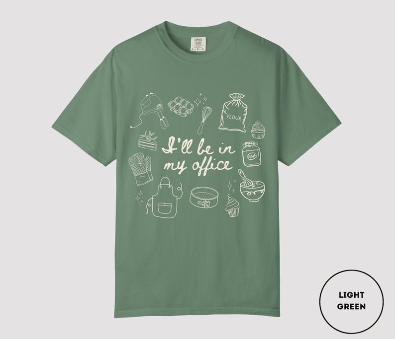 May include: Light green t-shirt with white line art of baking tools and the phrase "I'll be in my office." The design includes a mixer, whisk, oven mitt, apron, and cupcakes. The shirt is a comfortable, casual style.