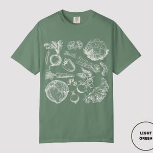 May include: Light green t-shirt featuring a detailed white illustration of various vegetables, including a beet, pepper, carrot, and cabbage. The shirt is a solid color with a crew neck and short sleeves. The text "LIGHT GREEN" is in a circle in the bottom right corner.