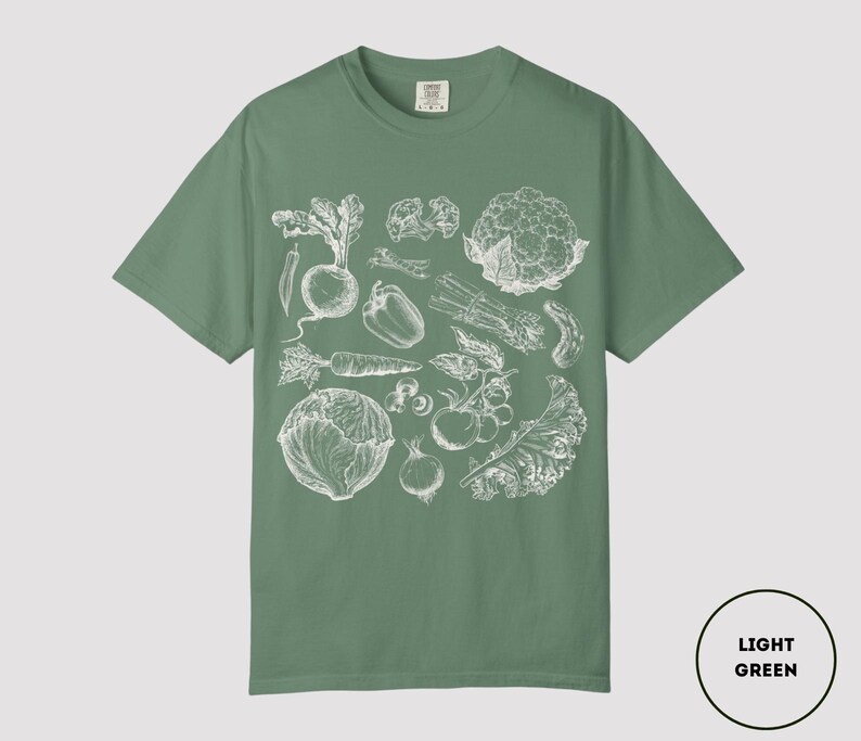 May include: Light green t-shirt featuring a detailed white illustration of various vegetables, including a beet, pepper, carrot, and cabbage. The shirt is a solid color with a crew neck and short sleeves. The text "LIGHT GREEN" is in a circle in the bottom right corner.