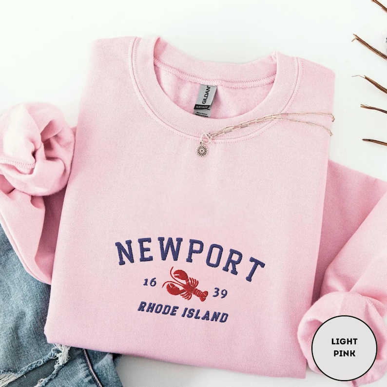 May include: A pale pink sweatshirt featuring the word "NEWPORT" in navy blue, with the numbers "16" and "39", and a red lobster graphic. The text "RHODE ISLAND" is below. A delicate necklace with a small pendant rests on the sweatshirt.