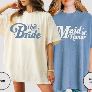 May include: Two oversized t-shirts are shown, one ivory and one blue. The ivory shirt features "the Bride" in blue script, while the blue shirt says "Maid of Honor" in white. Both are short-sleeved and have a retro design, ideal for a bridal party.
