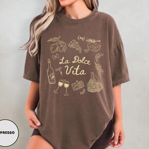 May include: Espresso-colored t-shirt with a gold graphic of Italian food and drink, including lemons, pizza, wine, and the phrase "La Dolce Vita". The shirt has a relaxed fit and short sleeves.
