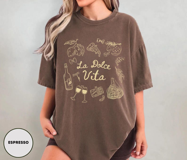 May include: Espresso-colored t-shirt with a gold graphic of Italian food and drink, including lemons, pizza, wine, and the phrase "La Dolce Vita". The shirt has a relaxed fit and short sleeves.