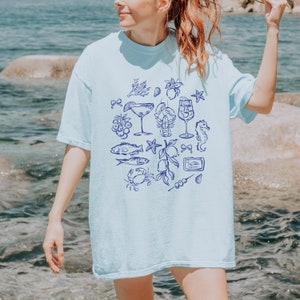 May include: Light blue t-shirt featuring a navy blue illustration of seafood and cocktails. The design includes images of a lobster, fish, seahorse, and martini glass. The shirt is a comfortable, casual style.