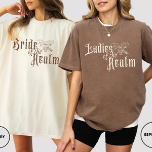 May include: Two oversized t-shirts, one ivory and one espresso, featuring the text "Bride of the Realm" and "Ladies of the Realm" in a decorative font. The shirts have a relaxed fit and short sleeves.