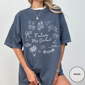 May include: Denim blue oversized t-shirt with white graphic illustrations of wedding-related items, including champagne glasses, shoes, and a bouquet. The text "Future Mrs. Earhart" is also printed on the front.