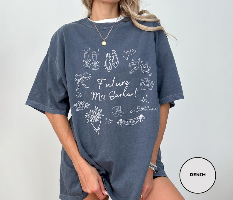 May include: Denim blue oversized t-shirt with white graphic illustrations of wedding-related items, including champagne glasses, shoes, and a bouquet. The text "Future Mrs. Earhart" is also printed on the front.