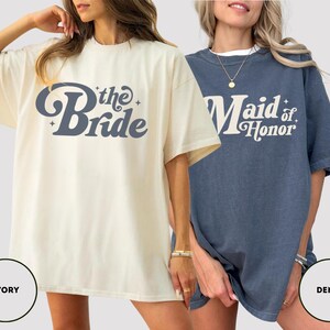 May include: Two oversized t-shirts, one ivory and one denim blue. The ivory shirt has "the Bride" in a retro font. The denim shirt says "Maid of Honor" in a similar style. Both are short-sleeved and suitable for a bridal party.