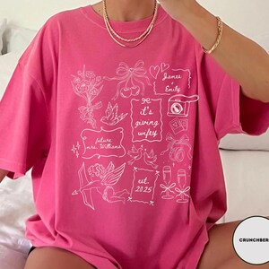 May include: A bright pink t-shirt with white line art illustrations of wedding-themed elements, including flowers, bows, doves, and champagne glasses. The text "it's giving wifey" is featured, along with names and the year "est. 2025".