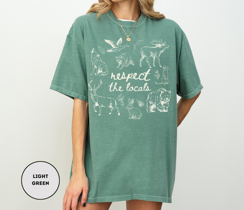 May include: Light green t-shirt with a crew neck and short sleeves. The shirt features a white graphic of various animals and the text "respect the locals". The shirt is made of a soft, comfortable material.