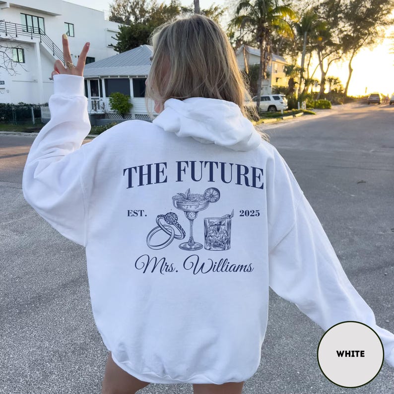 Pode incluir: Moletom branco com o texto "The Future Est. 2025 Mrs. Williams" estampado nas costas. O design inclui um copo de coquetel, um copo de margarita e duas alian&ccedil;as.