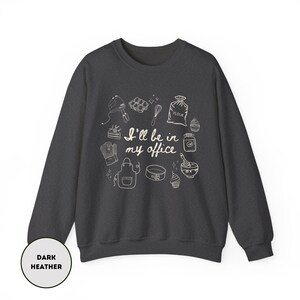 May include: Dark heather gray sweatshirt with a white graphic of baking tools and the text "I'll be in my office." The design includes a mixer, whisk, oven mitt, apron, and other kitchen items. A round label at the bottom left says "Dark Heather."