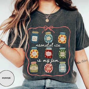 May include: Dark gray t-shirt with a pink outline featuring illustrations of jam jars. The jars have various fruit designs and text that reads "neonatal icu is my jam". The shirt has a crew neck and short sleeves.