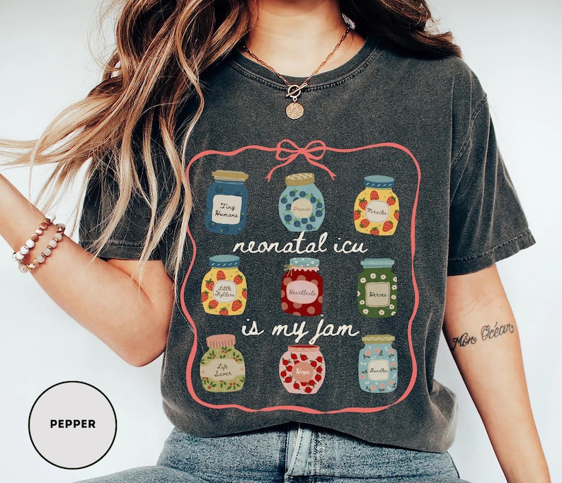 May include: Dark gray t-shirt with a pink outline featuring illustrations of jam jars. The jars have various fruit designs and text that reads "neonatal icu is my jam". The shirt has a crew neck and short sleeves.