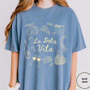 May include: Blue jean-colored t-shirt with a yellow graphic design. The design includes illustrations of lemons, pizza, wine glasses, and the text "La Dolce Vita". The shirt is a comfortable, casual style.