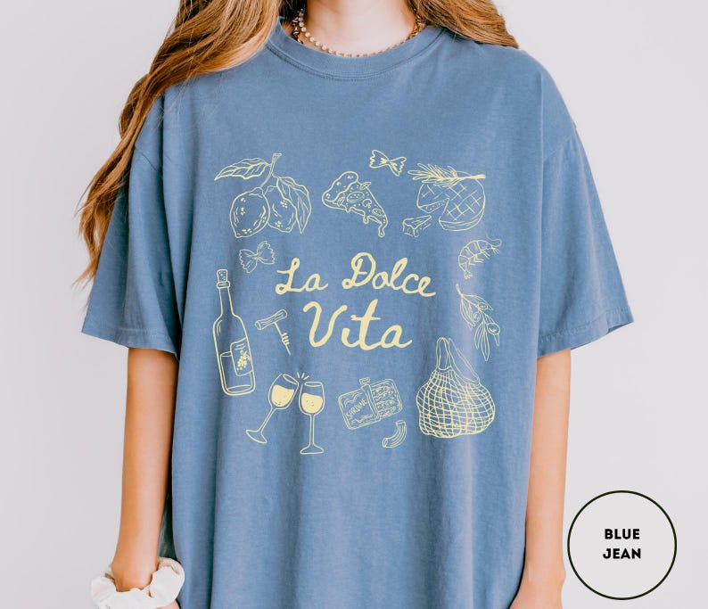 May include: Blue jean-colored t-shirt with a yellow graphic design. The design includes illustrations of lemons, pizza, wine glasses, and the text "La Dolce Vita". The shirt is a comfortable, casual style.