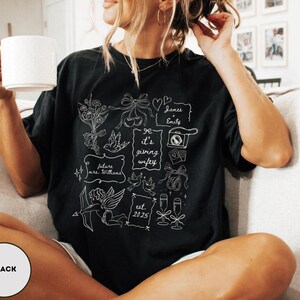 May include: Black t-shirt with a white graphic design featuring wedding-themed illustrations, including flowers, bows, doves, and the text "It's giving wifey." Additional text includes "future mrs. Williams" and "est. 2025."