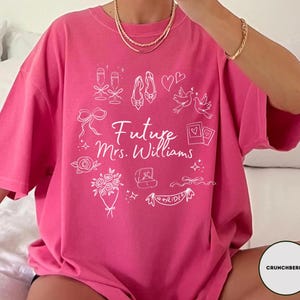 May include: A bright pink t-shirt with the text "Future Mrs. Williams" in white script. The shirt features white line drawings of wedding-related icons, including champagne glasses, shoes, and a ring.