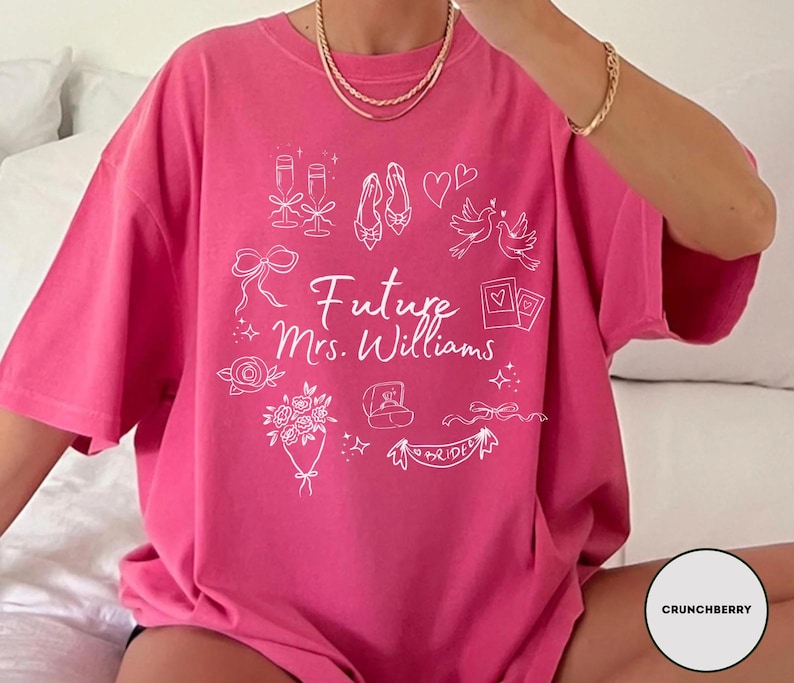 May include: A bright pink t-shirt with the text "Future Mrs. Williams" in white script. The shirt features white line drawings of wedding-related icons, including champagne glasses, shoes, and a ring.