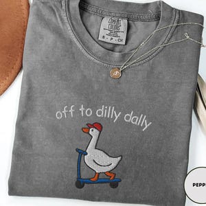May include: A gray Comfort Colors t-shirt with the embroidered phrase "off to dilly dally" and a cartoon goose on a scooter. The goose wears a red cap. A silver necklace with a pendant is draped on the shirt.