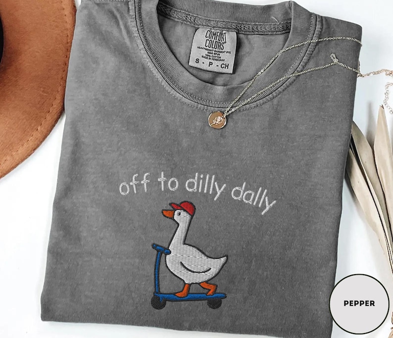 May include: A gray Comfort Colors t-shirt with the embroidered phrase "off to dilly dally" and a cartoon goose on a scooter. The goose wears a red cap. A silver necklace with a pendant is draped on the shirt.