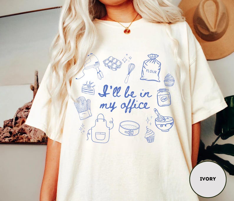 May include: Ivory-colored t-shirt with blue line art illustrations of baking tools and ingredients. The text "I'll be in my office" is printed in blue. The shirt features a relaxed fit and short sleeves.