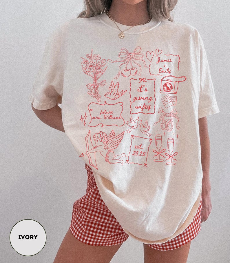 May include: Ivory-colored t-shirt with red graphic illustrations, including flowers, bows, a camera, and text that reads "James + Emily", "It's giving wifey", "future mrs. Williams", and "est. 2025".