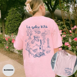 Puede incluir: Una camiseta rosa con un estampado gr&aacute;fico azul de varios alimentos italianos, incluyendo una langosta, uvas, ostras, una botella de vino y un sacacorchos. El texto "La Dolce Vita" est&aacute; impreso en la camiseta.