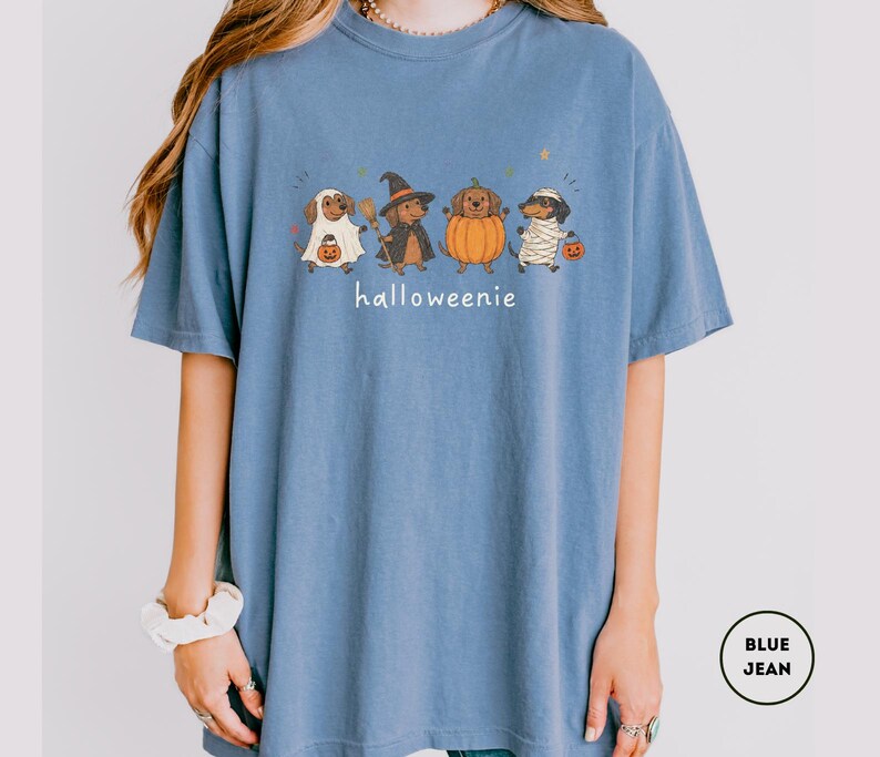 Op de afbeelding: Een blauw spijkerkleurig t-shirt met een afbeelding van vier teckels verkleed in Halloween-kostuums als geest, heks, pompoen en mummie. De tekst "halloweenie" staat in het wit onder de honden gedrukt.