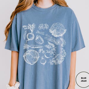 May include: A blue jean-colored t-shirt featuring a detailed white illustration of various vegetables, including a beet, carrot, and cauliflower. The design is centered on the shirt, showcasing a botanical theme. The shirt is made of a soft material.