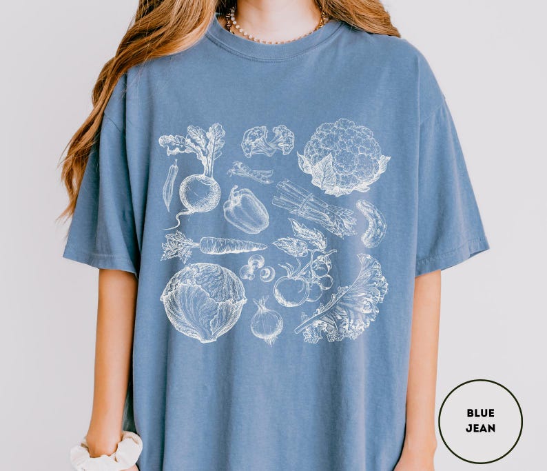 May include: A blue jean-colored t-shirt featuring a detailed white illustration of various vegetables, including a beet, carrot, and cauliflower. The design is centered on the shirt, showcasing a botanical theme. The shirt is made of a soft material.