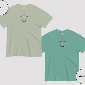 May include: Two short-sleeved t-shirts, one in bay green and the other in seafoam green. Each shirt features an embroidered design of a goose on a scooter with the text "off to dilly daily" above it. The shirts are made of a soft material.