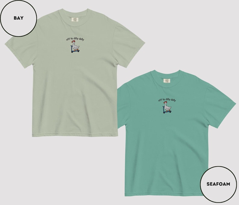 May include: Two short-sleeved t-shirts, one in bay green and the other in seafoam green. Each shirt features an embroidered design of a goose on a scooter with the text "off to dilly daily" above it. The shirts are made of a soft material.