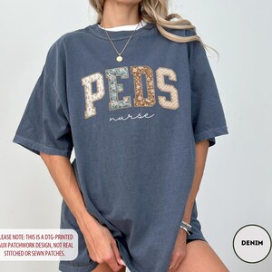 May include: A denim blue t-shirt with the word "PEDS" in a faux patchwork design, with the word "nurse" underneath. The shirt has short sleeves and a relaxed fit. The text "PLEASE NOTE: THIS IS A DTG-PRINTED FAUX PATCHWORK DESIGN, NOT REAL STITCHED OR SEWN PATCHES." is at the bottom.