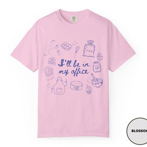 May include: A light pink t-shirt with a blue illustration of baking tools and the phrase "I'll be in my office." The design includes a mixer, eggs, a whisk, a bag of flour, a cupcake, and more. The shirt is labeled "Blossom."