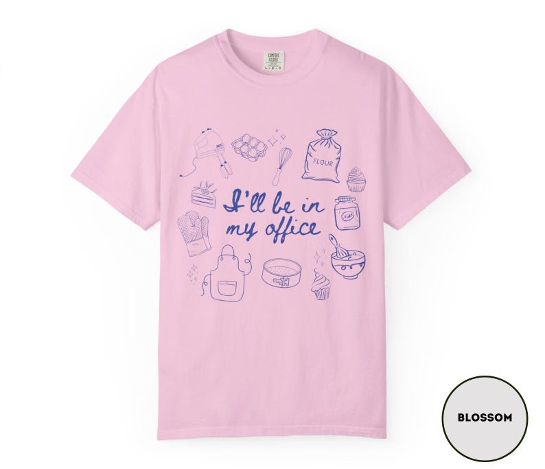 May include: A light pink t-shirt with a blue illustration of baking tools and the phrase "I'll be in my office." The design includes a mixer, eggs, a whisk, a bag of flour, a cupcake, and more. The shirt is labeled "Blossom."
