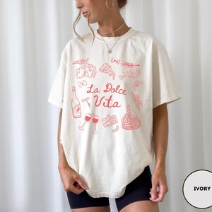 May include: Ivory-colored t-shirt with a red graphic design featuring Italian food and drink illustrations, including a wine bottle, glasses, and pizza. The words "La Dolce Vita" are written in a script font.