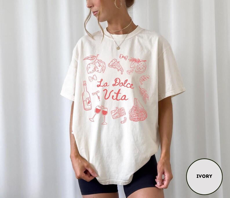 May include: Ivory-colored t-shirt with a red graphic design featuring Italian food and drink illustrations, including a wine bottle, glasses, and pizza. The words "La Dolce Vita" are written in a script font.