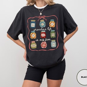 Peut inclure: T-shirt noir avec un motif de neuf pots de confiture dans un cadre rose. Le texte "grandma life is my jam" est &eacute;crit en police script. Le t-shirt est de coupe d&eacute;contract&eacute;e et &agrave; manches courtes.
